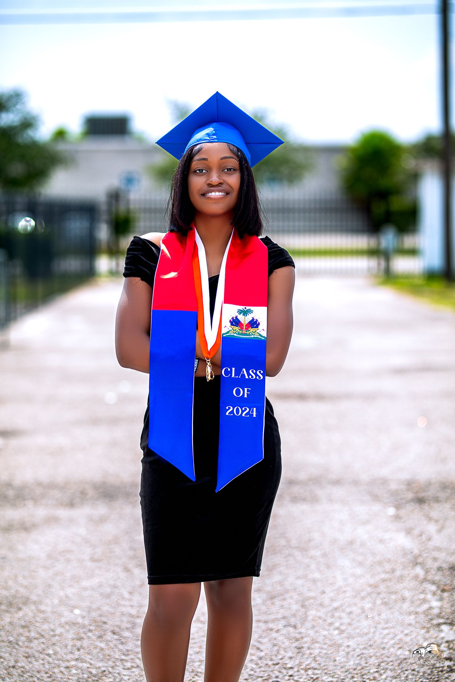 Graduate in blue cap and Haitian stole stands proudly with diploma outdoors.