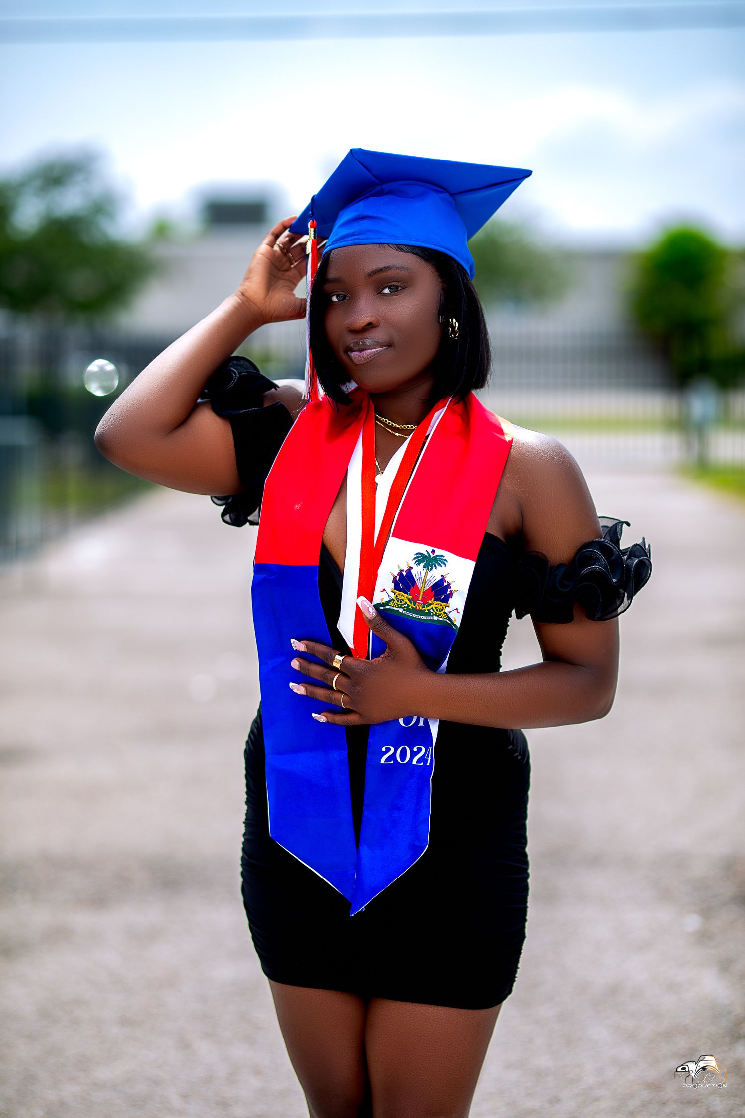 Graduate in blue cap and red Haitian stole celebrating academic achievement outdoors.