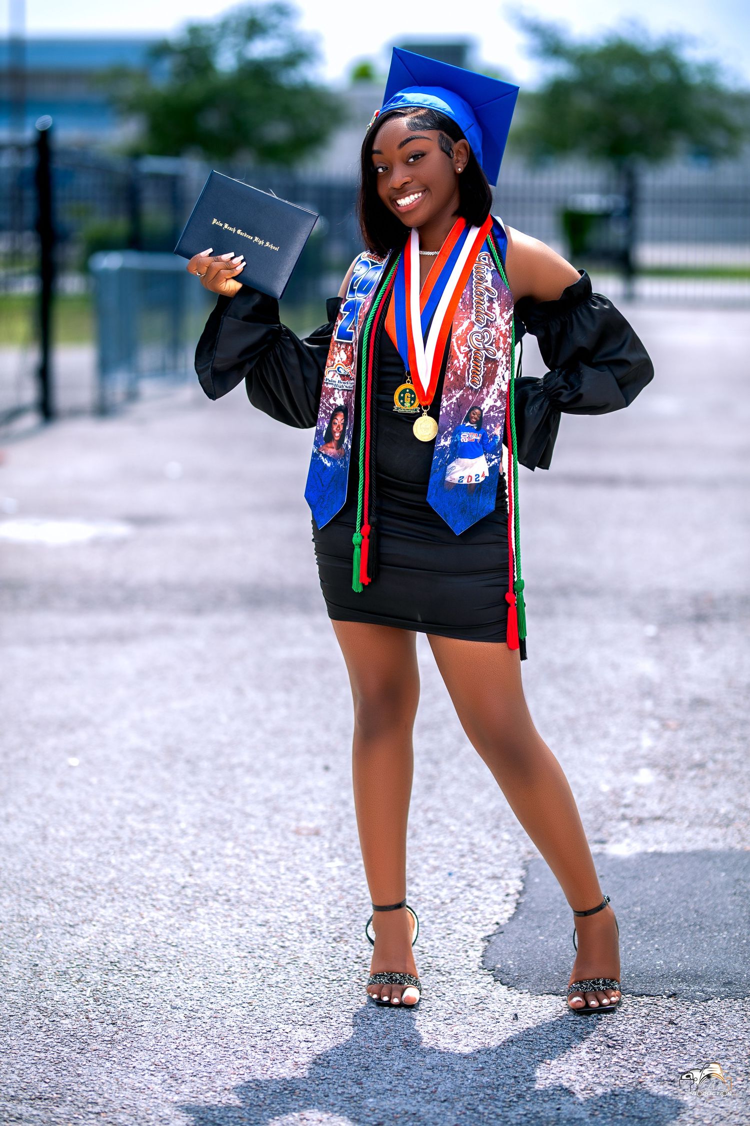 Graduate in blue cap and gown holding diploma with excited expression outdoors.