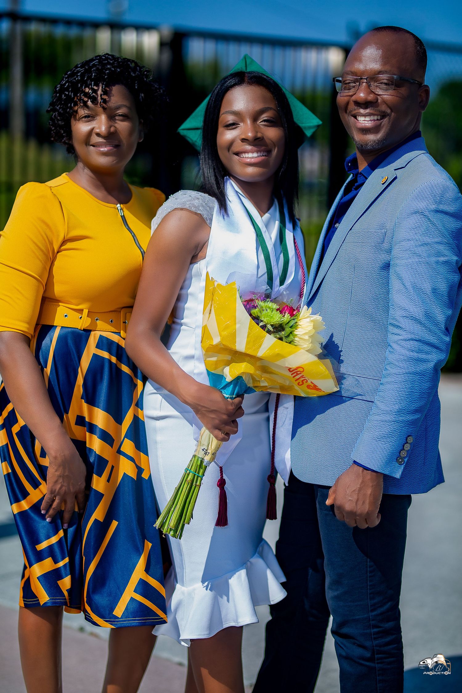 Graduate in white dress celebrating with family outdoors while holding yellow flower bouquet.