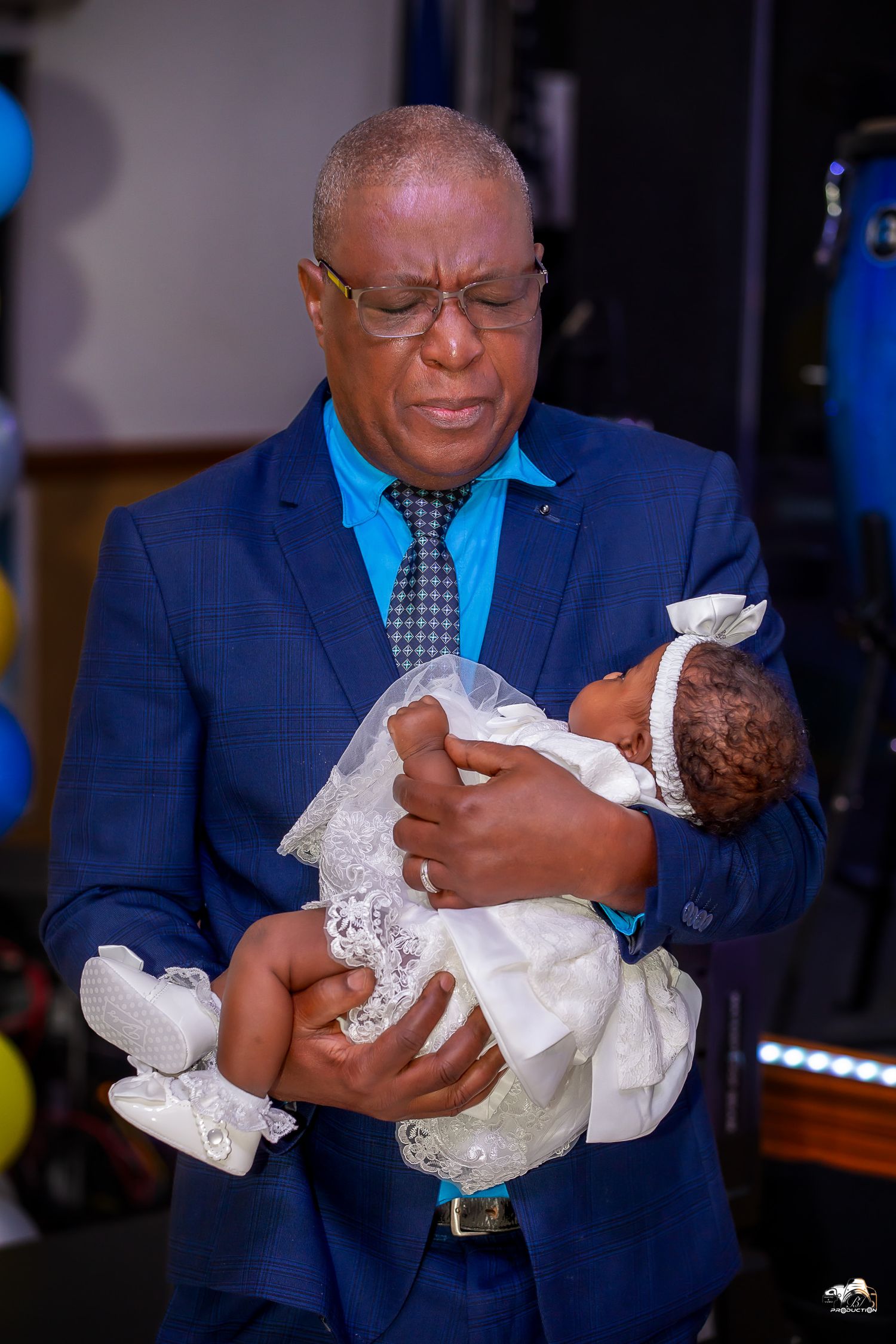 Elder in blue suit tenderly holds infant dressed in white baptismal gown.
