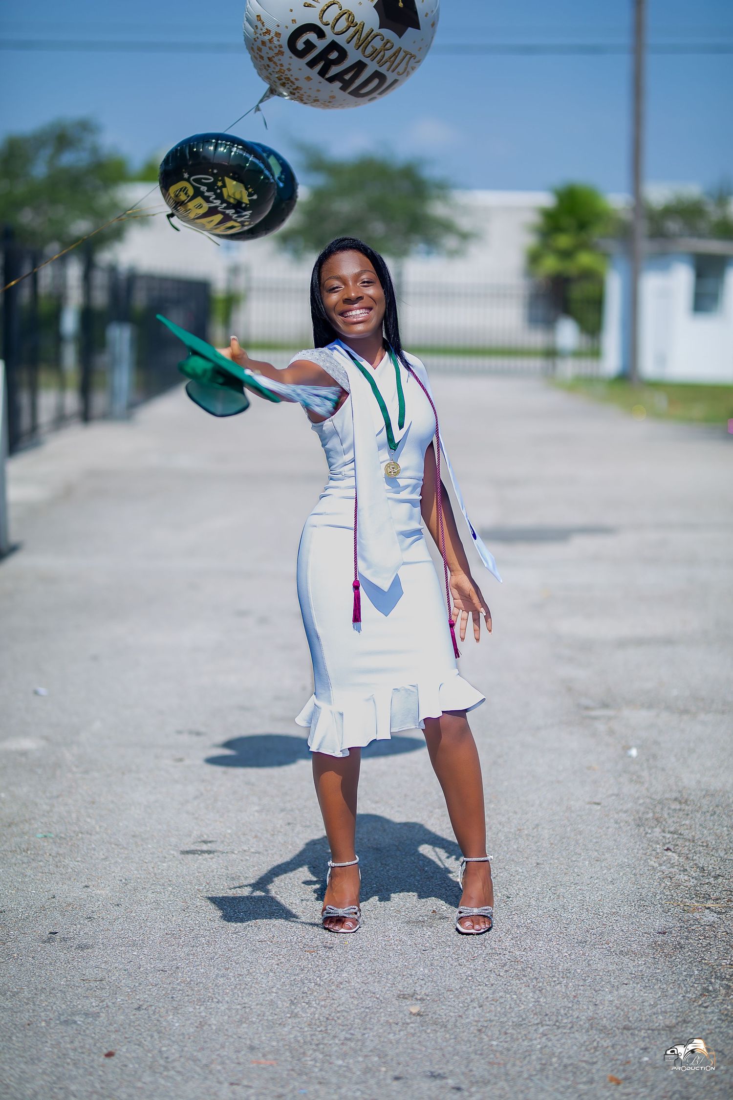 Graduate in white dress tossing celebration balloons in outdoor setting.