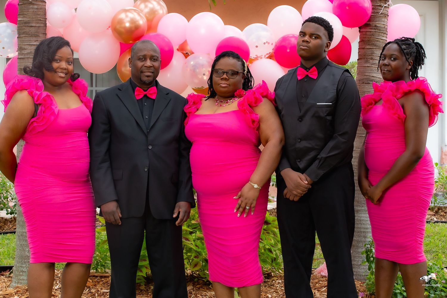 A group wearing coordinated bright pink dresses and black suits pose together in front of pink balloon decorations.