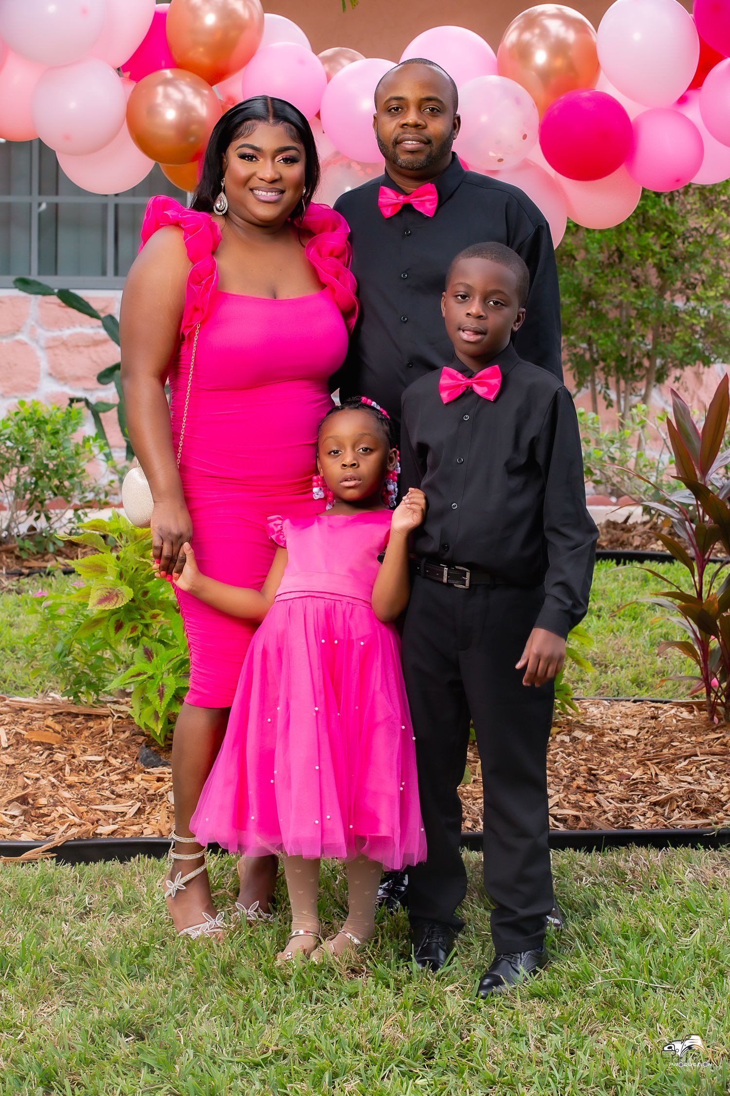 A family in coordinating pink and black formal attire poses against a pink balloon backdrop.