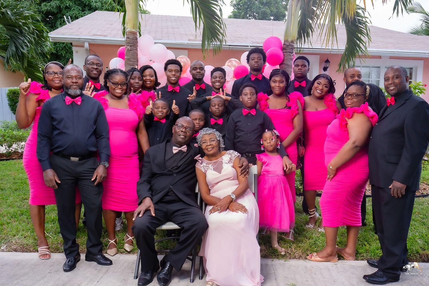 A large group in coordinating pink dresses and black suits poses outdoors for a formal celebration photo.