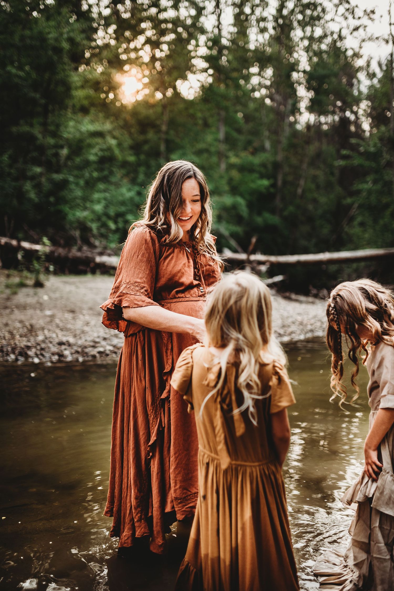 Three people wearing vintage-style dresses stand in a shallow creek at sunset with trees in the background, family photos
