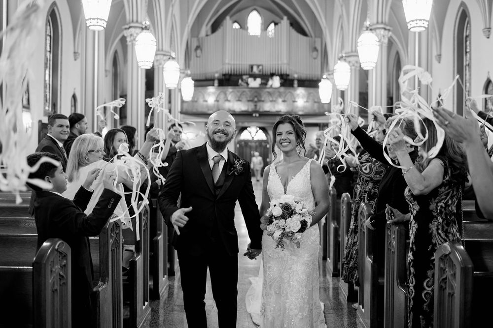 Bride and groom walk down church aisle as guests wave ribbon wands in black and white wedding photo.