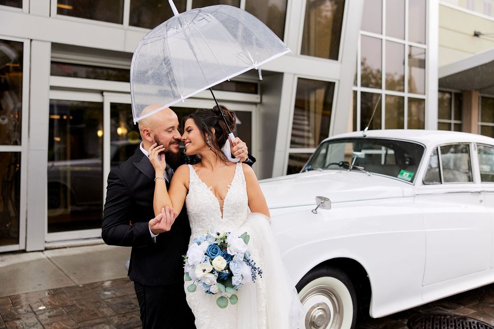 Bride and groom share a romantic moment under a clear umbrella beside a vintage white Rolls-Royce on a rainy wedding day.