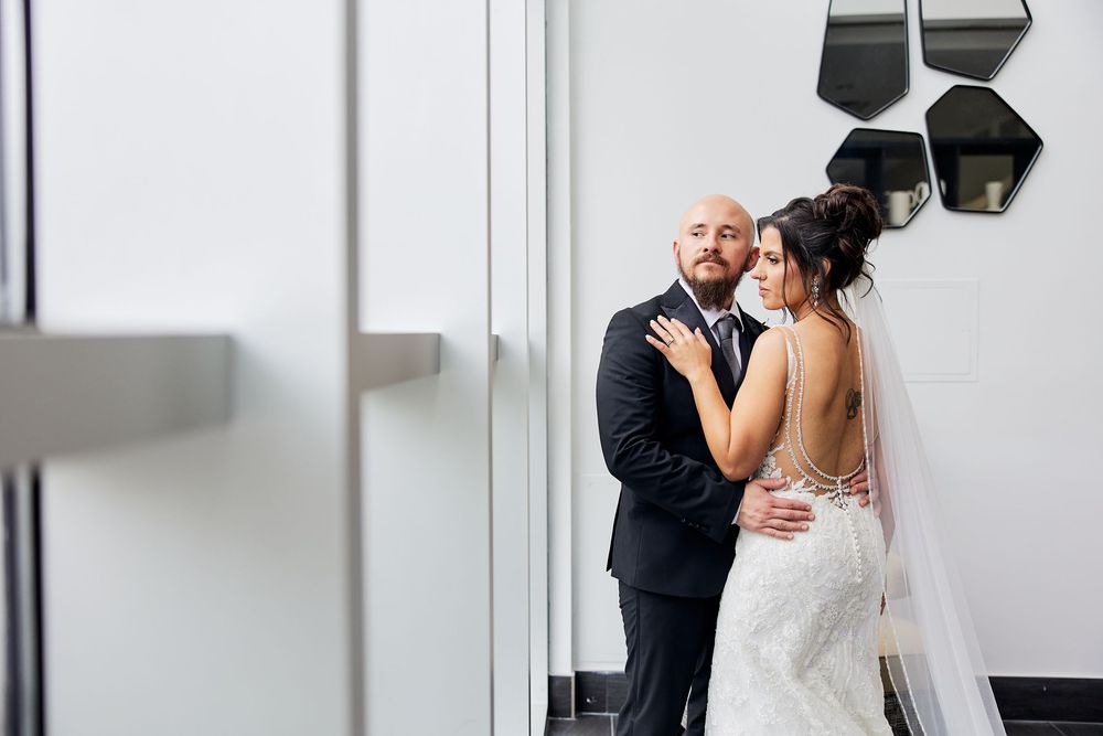 A bride and groom share a tender embrace in a modern white hallway, the bride wearing a lace gown and veil.