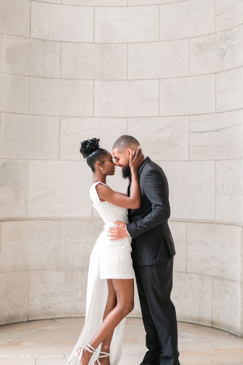 A couple in wedding attire shares a romantic embrace against a marble wall, the bride in white and groom in a black suit.