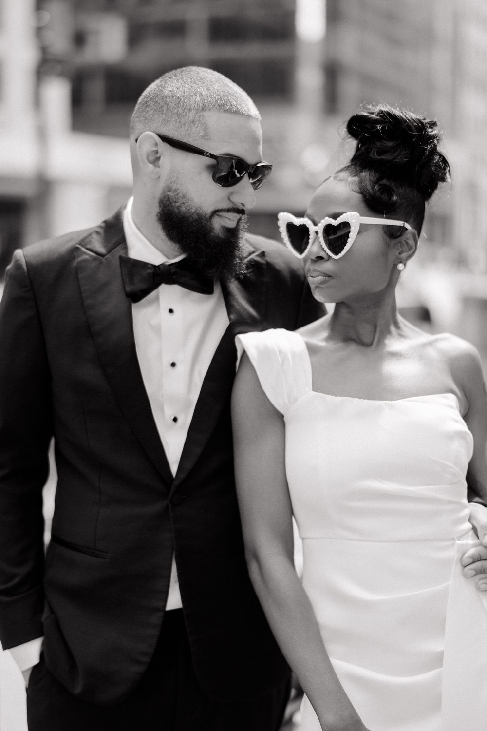 Black and white photo of a stylish couple in wedding attire, groom in tuxedo and bride in white dress with heart sunglasses.