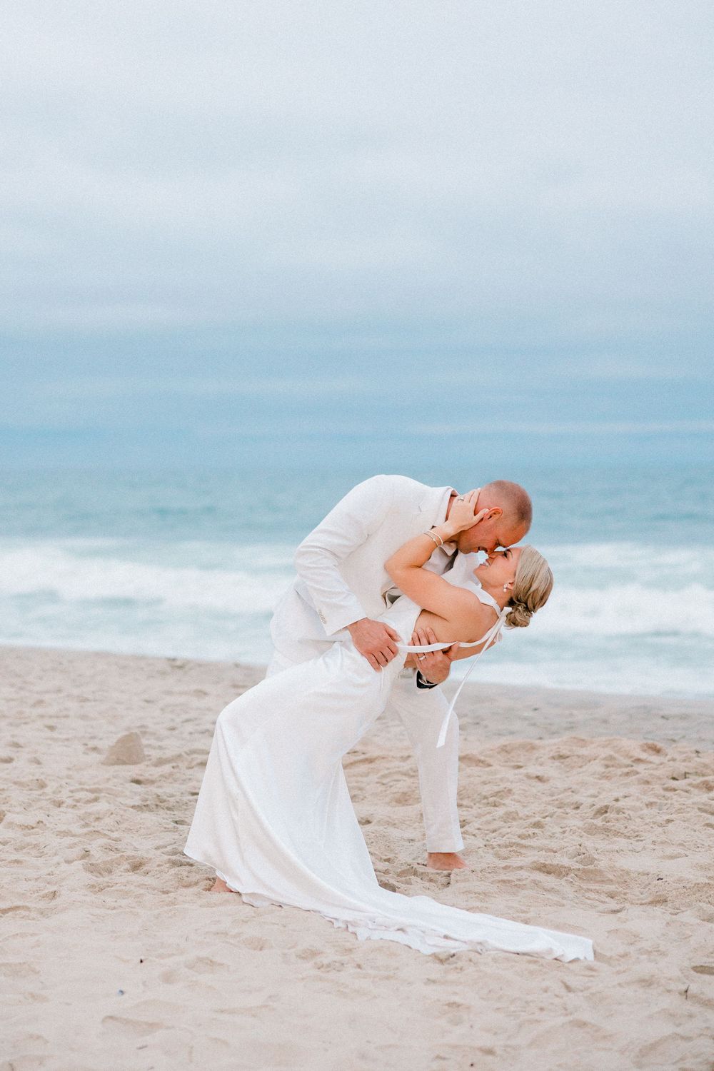 A couple in white attire performs a romantic dance dip pose on a serene beach with ocean waves in the background.