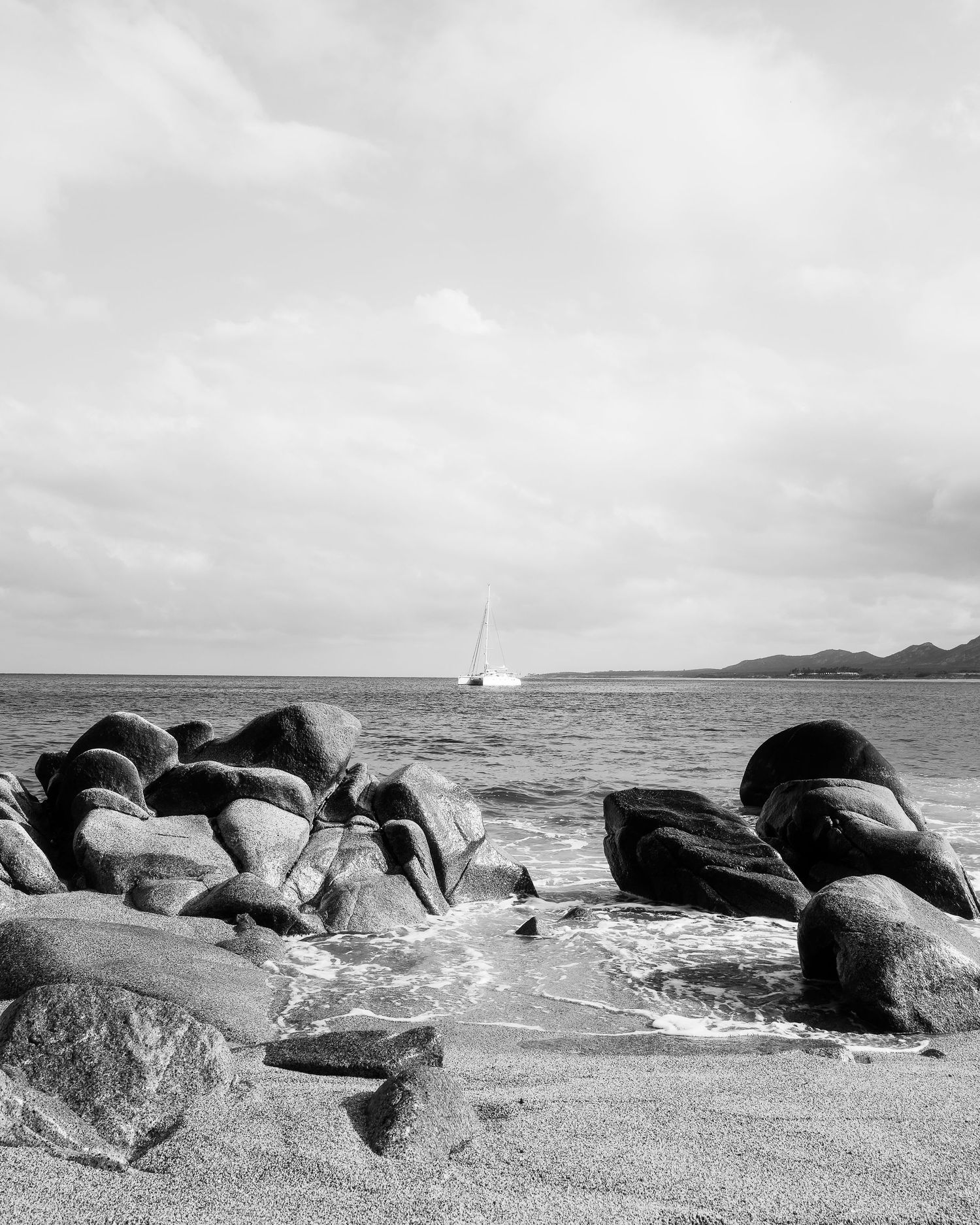 Black and white beach scene with large rocks, gentle waves, and a sailboat on calm ocean waters under cloudy skies.