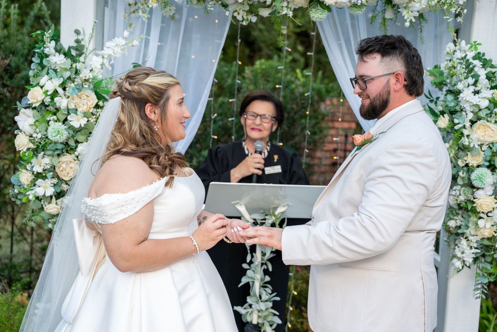 A wedding ceremony takes place under a floral arch with white curtains as a couple exchanges rings during their vows.