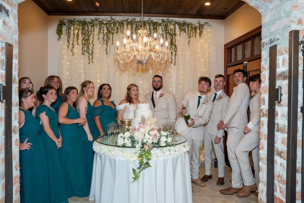 Wedding party in teal bridesmaid dresses and light suits gather around an elegant table with floral decor and chandelier.
