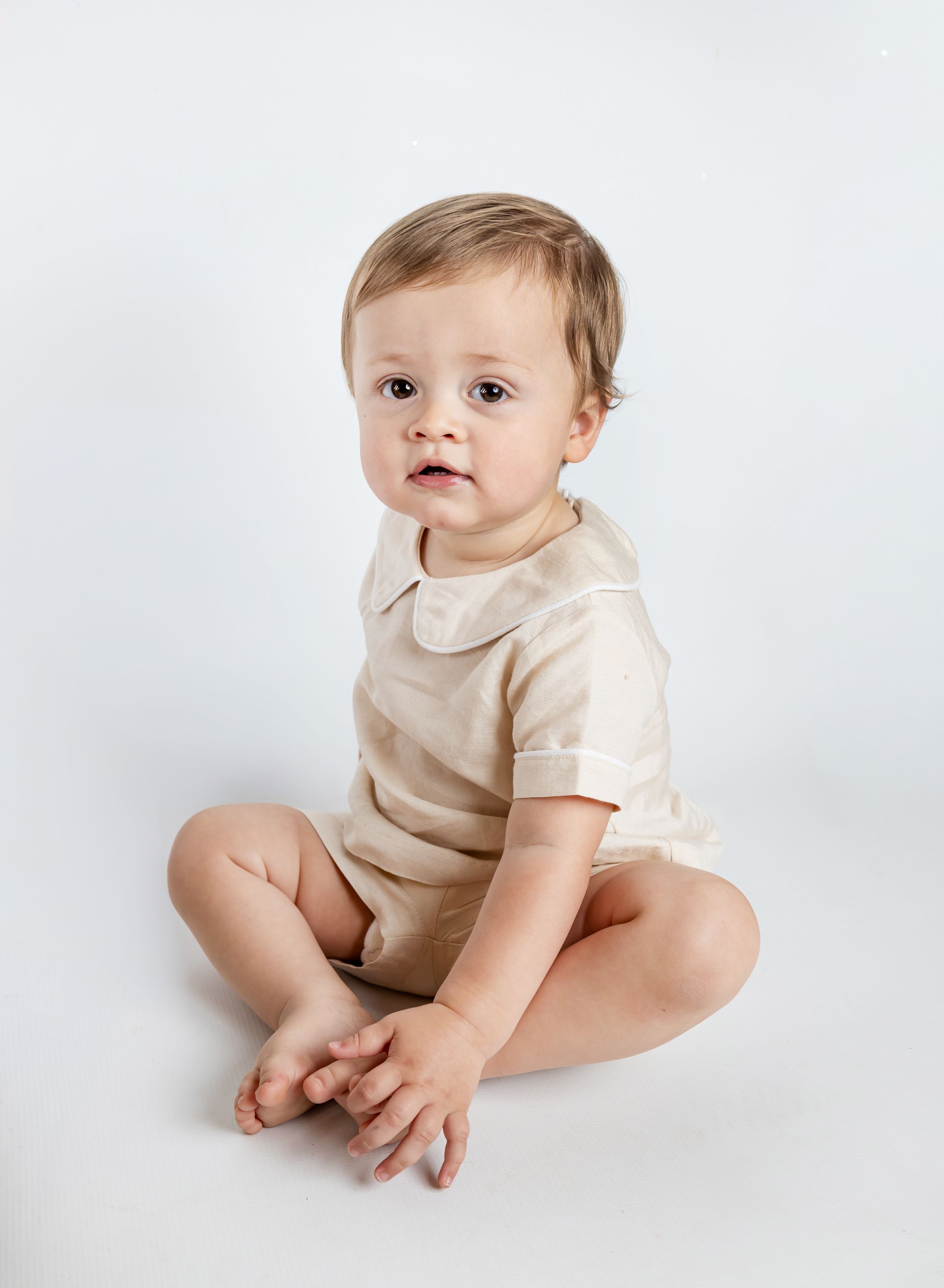 Baby wearing beige peter pan collar romper sits on white background with bare feet and gentle expression.