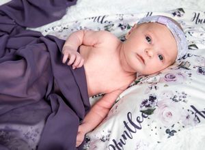 Baby wearing a blue hat lies on floral patterned bedding wrapped in a purple blanket.