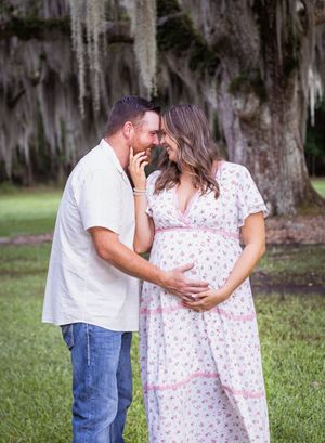 A couple shares a tender moment outdoors, the pregnant woman in a floral dress cradling her baby bump beneath moss-draped trees.