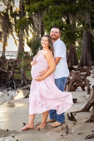 Pregnant couple posing on sandy beach, woman in pink maternity dress, man in white shirt and jeans, surrounded by cypress trees.