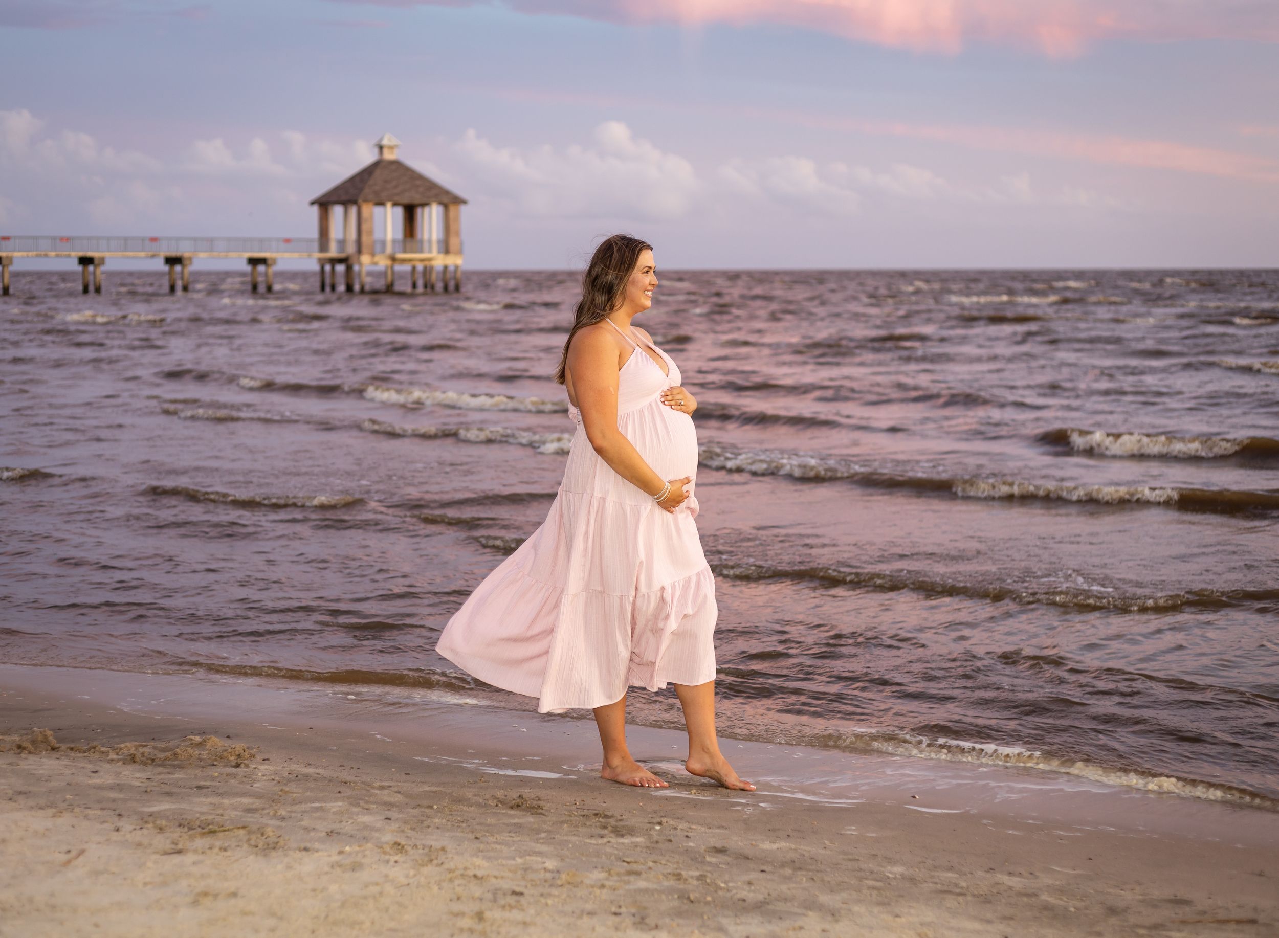 Pregnant woman in pink dress on beach at sunset with pier in background.