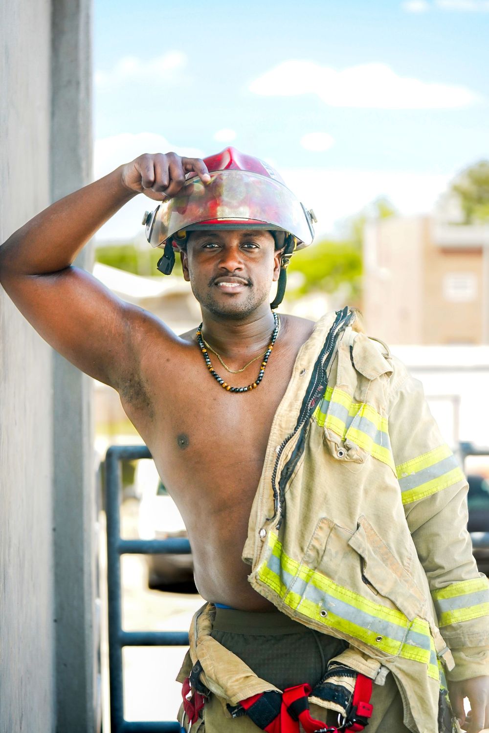 A shirtless Black male firefighter holds a red helmet while wearing open turnout gear outdoors on a sunny day.