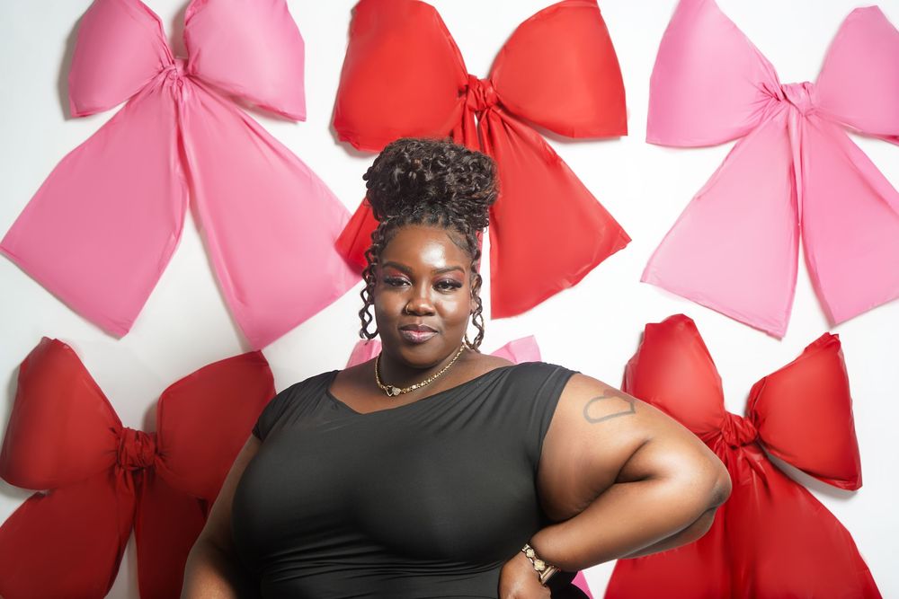 Woman in black dress poses confidently against white wall decorated with large red and pink bows.