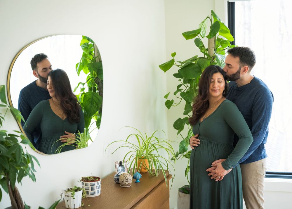 A couple embraces in front of a potted houseplant by a window in a bright, minimalist room.