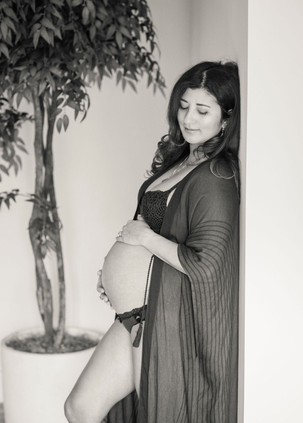 A black and white maternity photo showing an expectant mother in a long cardigan posing next to a potted plant.