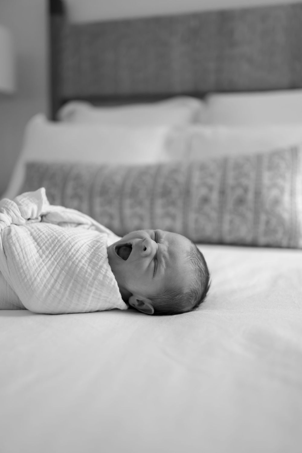 Black and white photo of a swaddled newborn baby yawning while lying on a white bed.