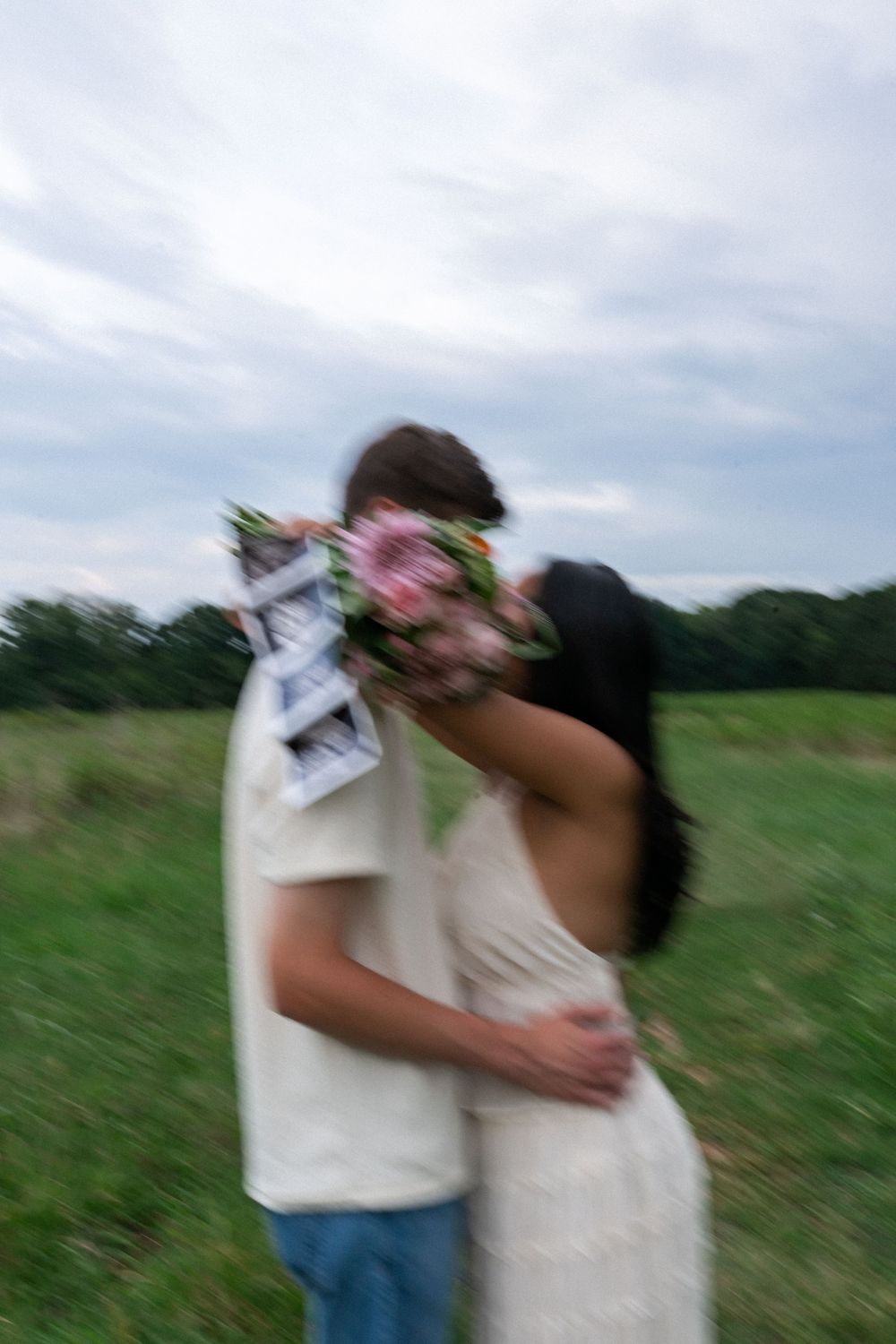 Couple kissing in a green field, woman in white dress holding pink flower bouquet, blurred romantic outdoor photo.