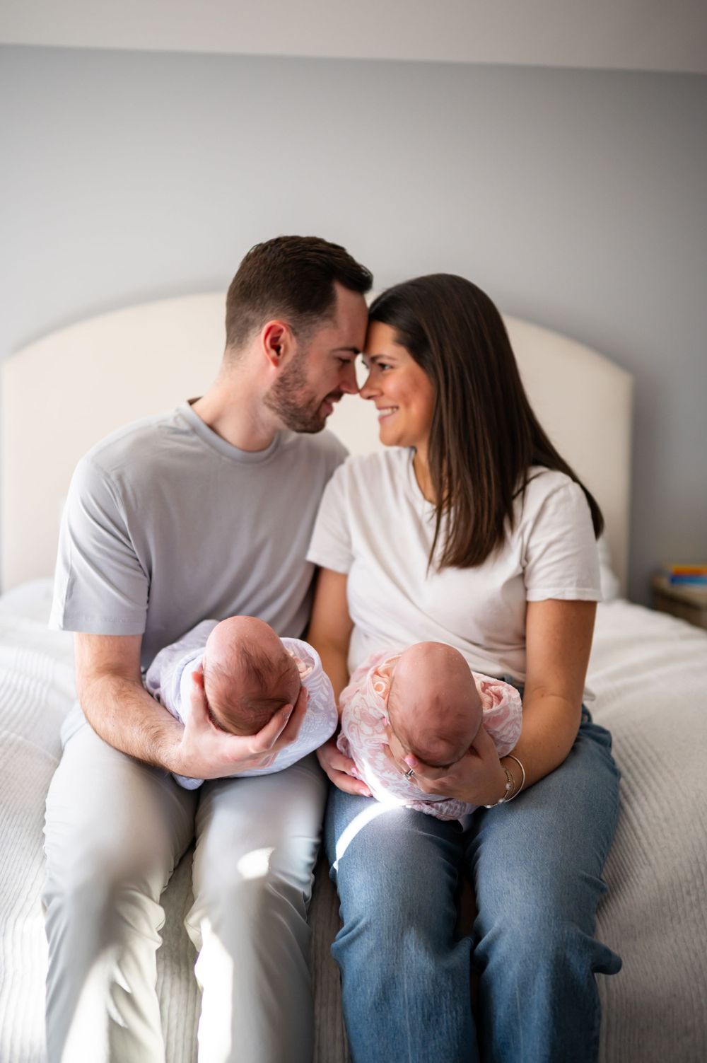Parents holding twin newborn babies while sitting together on a bed in a tender moment.