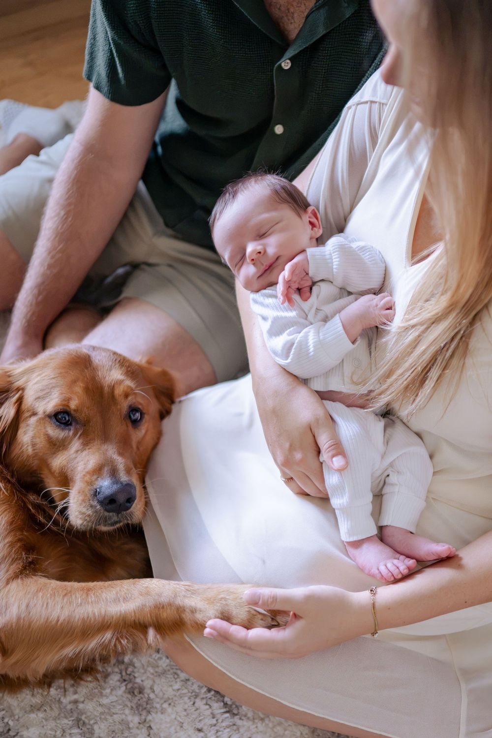 A golden retriever rests beside parents holding a sleeping newborn baby, with mom gently holding the dog's paw.