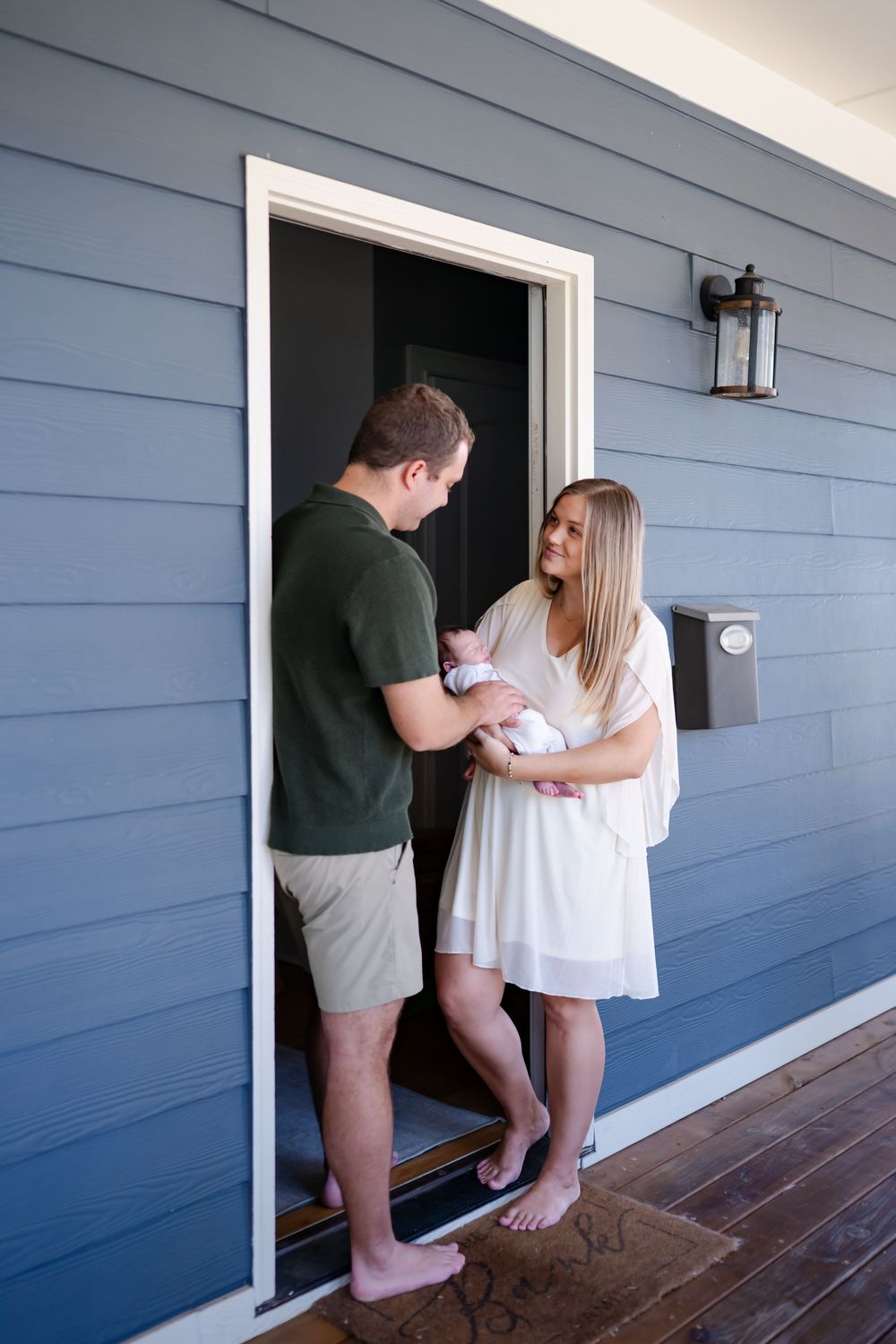 Young couple with newborn baby standing barefoot at front door of blue house.