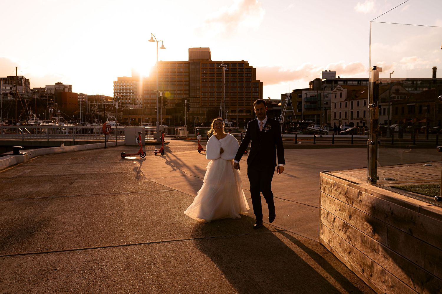 Wedding couple stroll an urban sidewalk during golden hour sunset along the Hobart waterfront. Gorgeous, natural wedding photography.