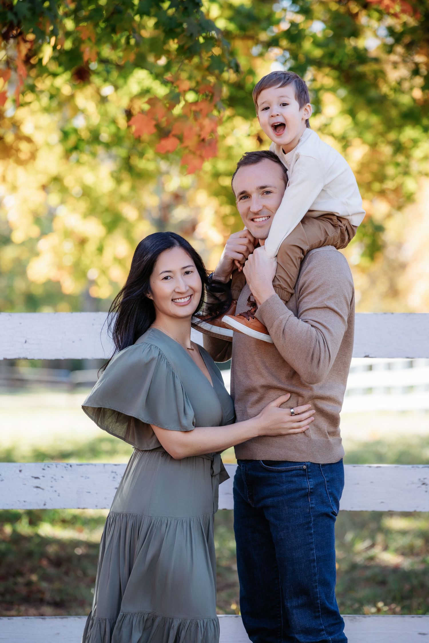A family poses together on an autumn day near a white fence with colorful fall foliage in the background.