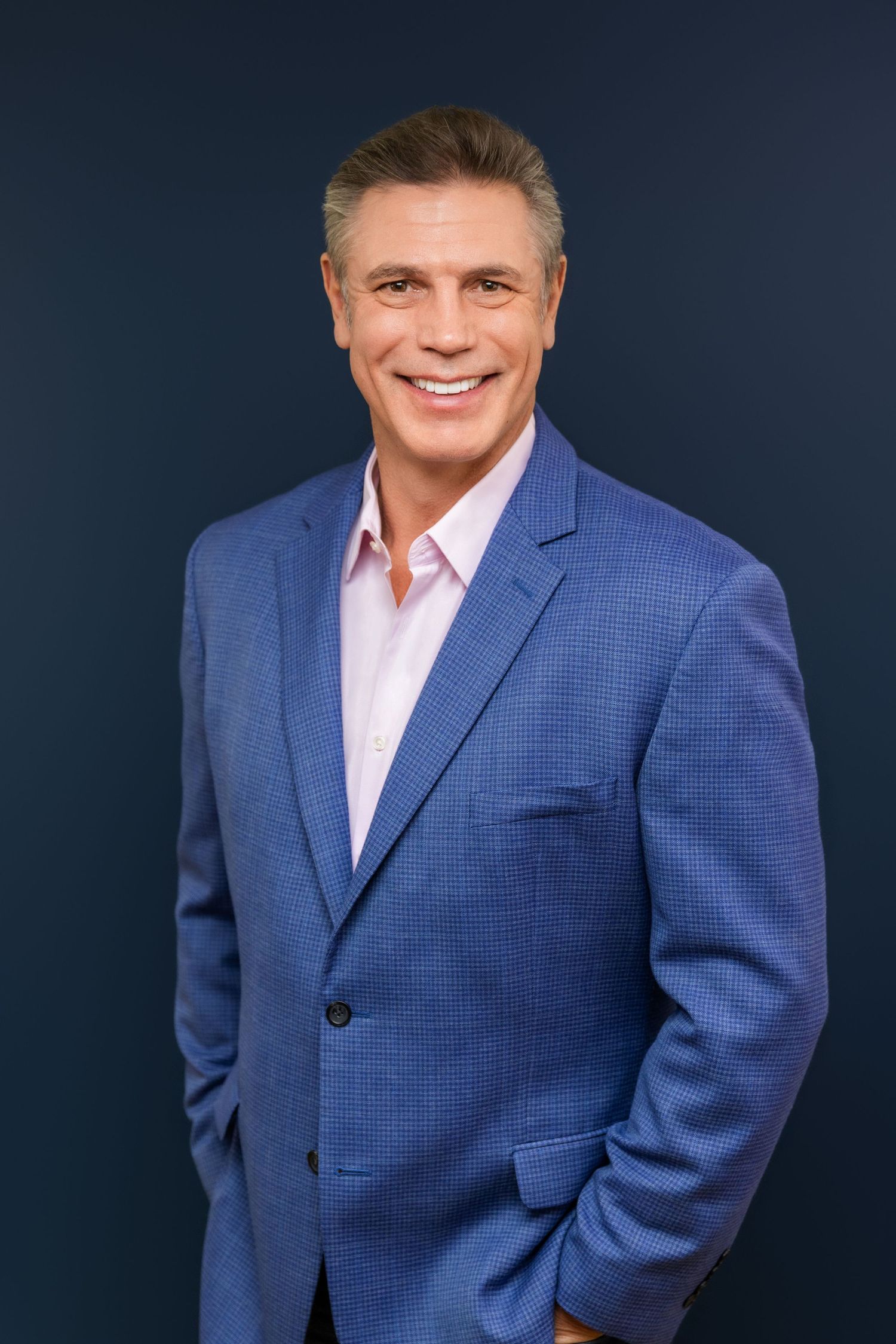 Professional headshot portrait of someone in a blue suit jacket and pink dress shirt against a dark background.
