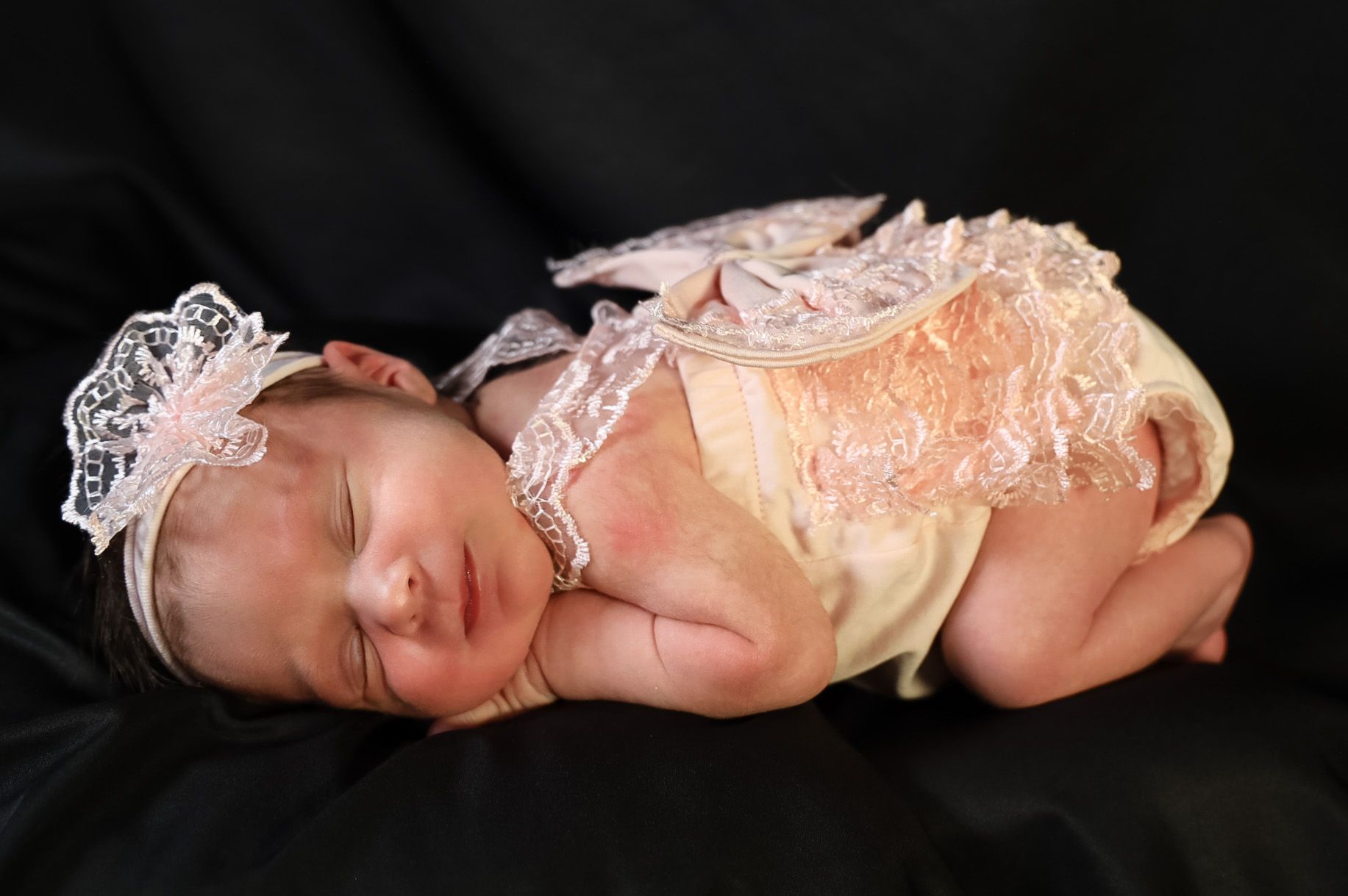 Sleeping newborn baby girl wearing lace headband and ruffled outfit on black background.