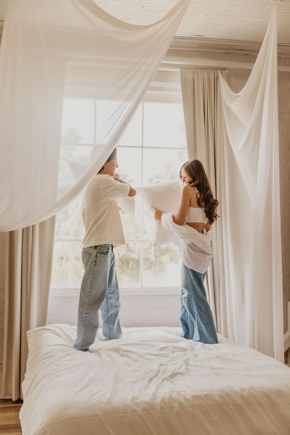 Two people in casual clothes put up light sheer curtains in a bright, airy bedroom with a white bed.