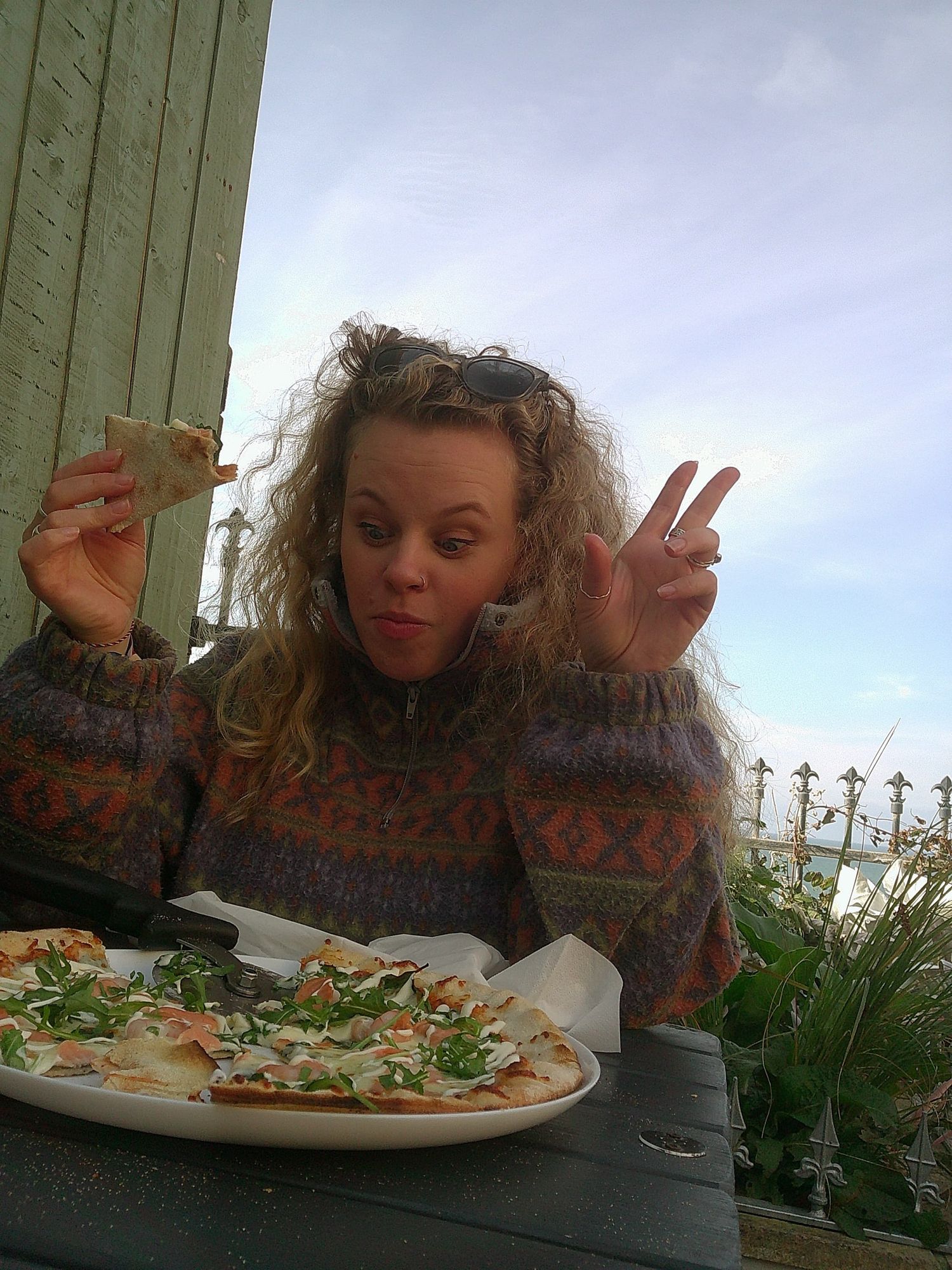 Young woman with curly hair making a peace sign while eating pizza outdoors, wearing a patterned sweater in Newquay