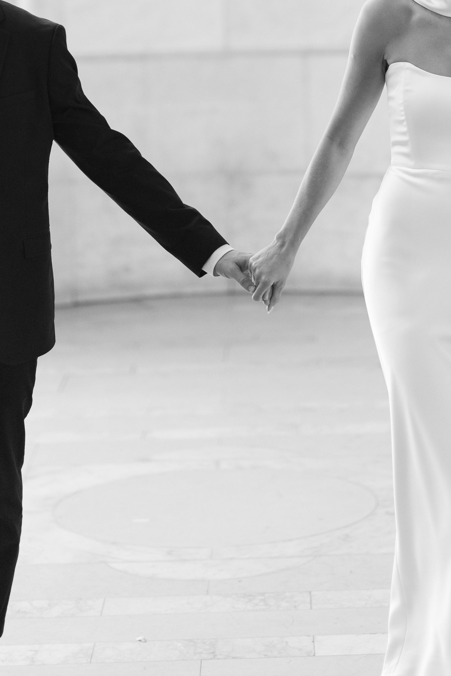 Black and white photo of a bride and groom holding hands in elegant wedding attire.