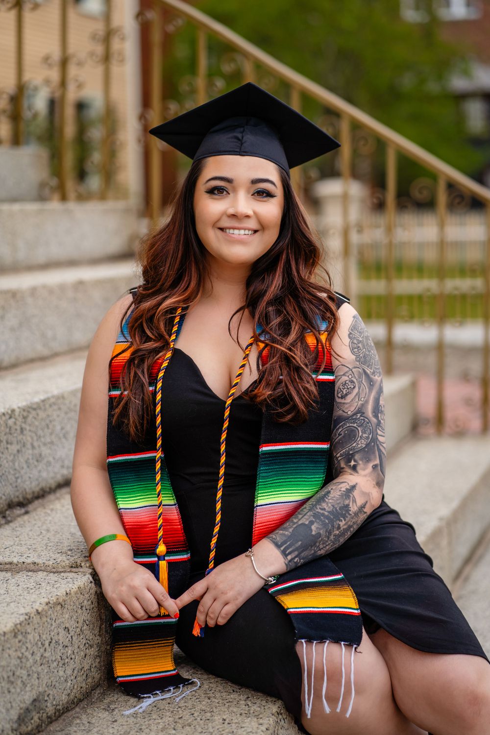 Graduate in traditional cap and gown with colorful serape stole sits on concrete steps outdoors.