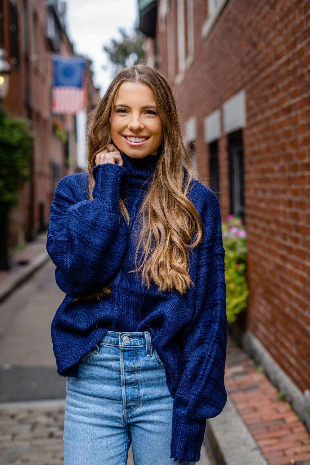 A casual street style portrait featuring a navy blue sweater and light wash jeans against a brick wall backdrop.