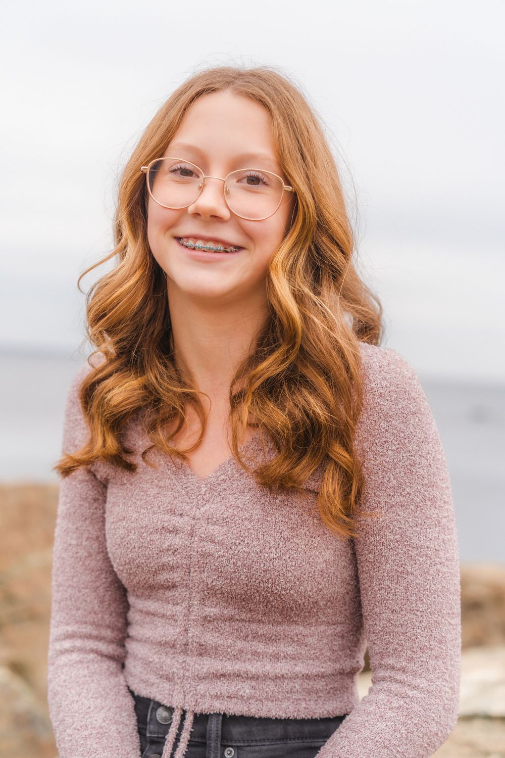 Portrait of person in pink sweater with glasses against cloudy coastal backdrop.