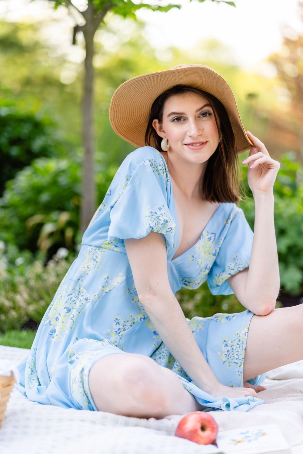 Woman in blue floral dress sits on blanket wearing straw hat in garden setting.