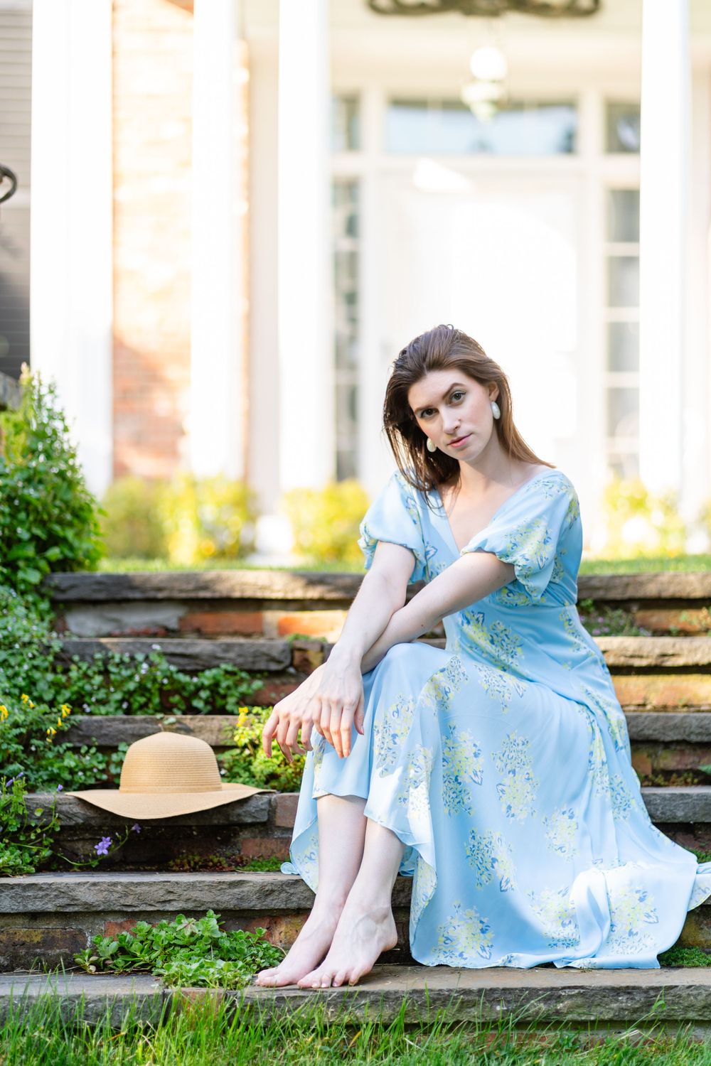 Woman in light blue dress poses barefoot on stone steps with straw hat beside her.