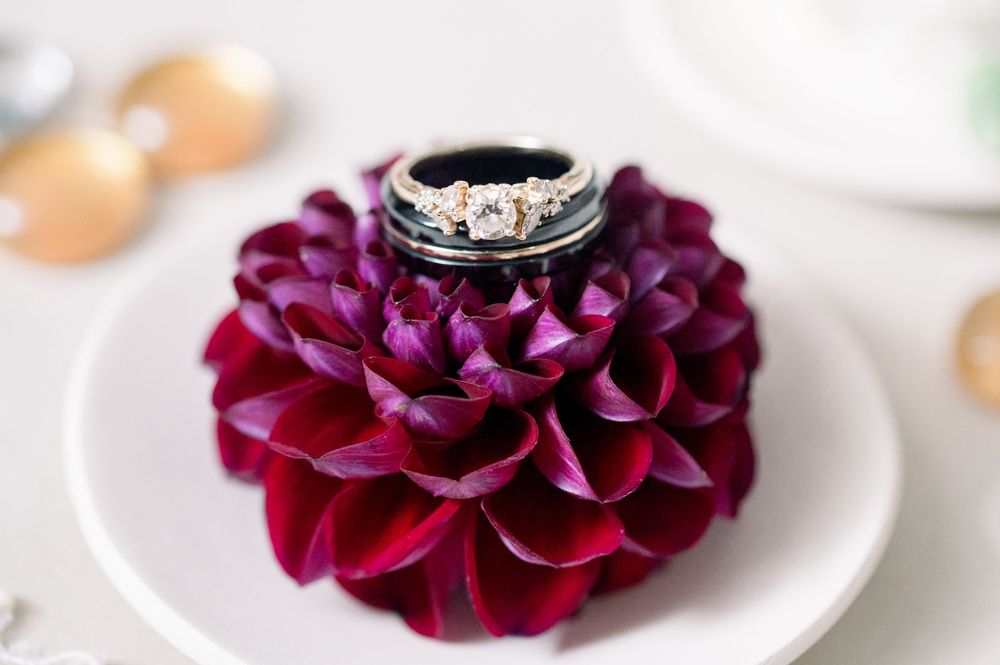 A deep burgundy dahlia flower with a diamond engagement ring nestled in its petals on a white plate.