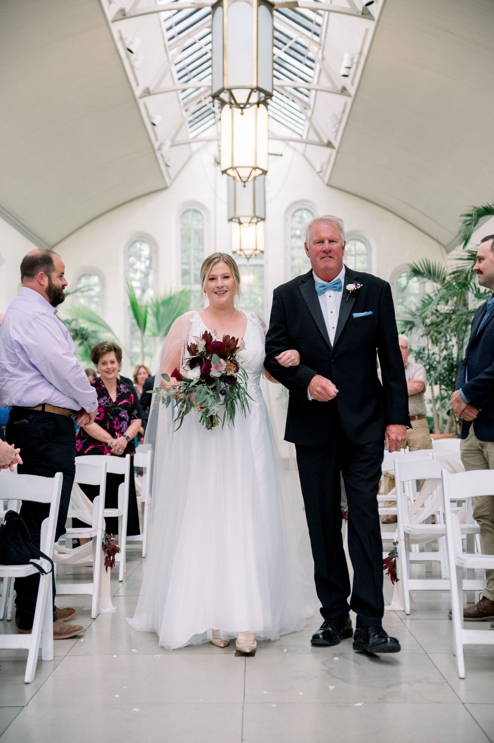 Wedding ceremony in a bright conservatory with vaulted glass ceiling and white chairs lining the aisle.