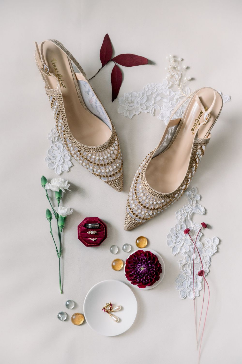 Elegant bridal accessories including pearl-adorned shoes, rings, and floral elements arranged on white lace background.