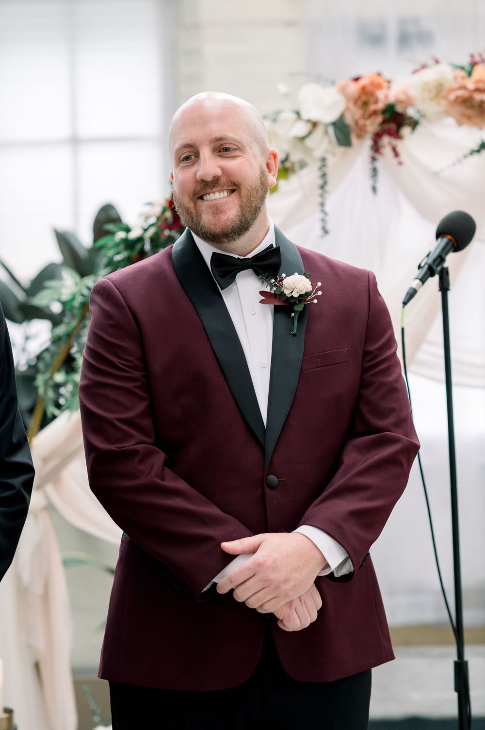 A groom in a burgundy tuxedo jacket and black bowtie smiles joyfully during a wedding celebration.
