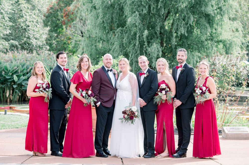 Wedding party poses together in formal attire with bridesmaids wearing bright red dresses in an outdoor garden setting.